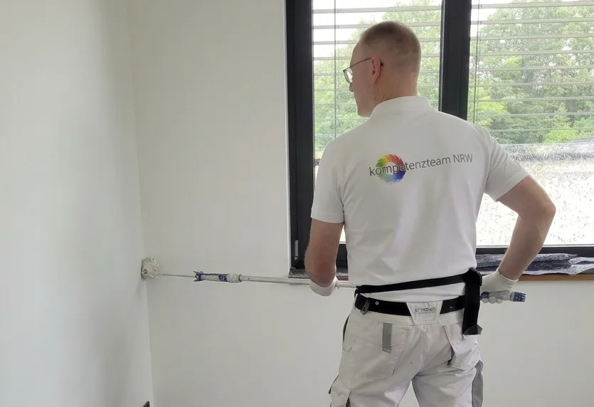 Maler von Kompetenzteam NRW streicht eine helle Innenwand am Fenster